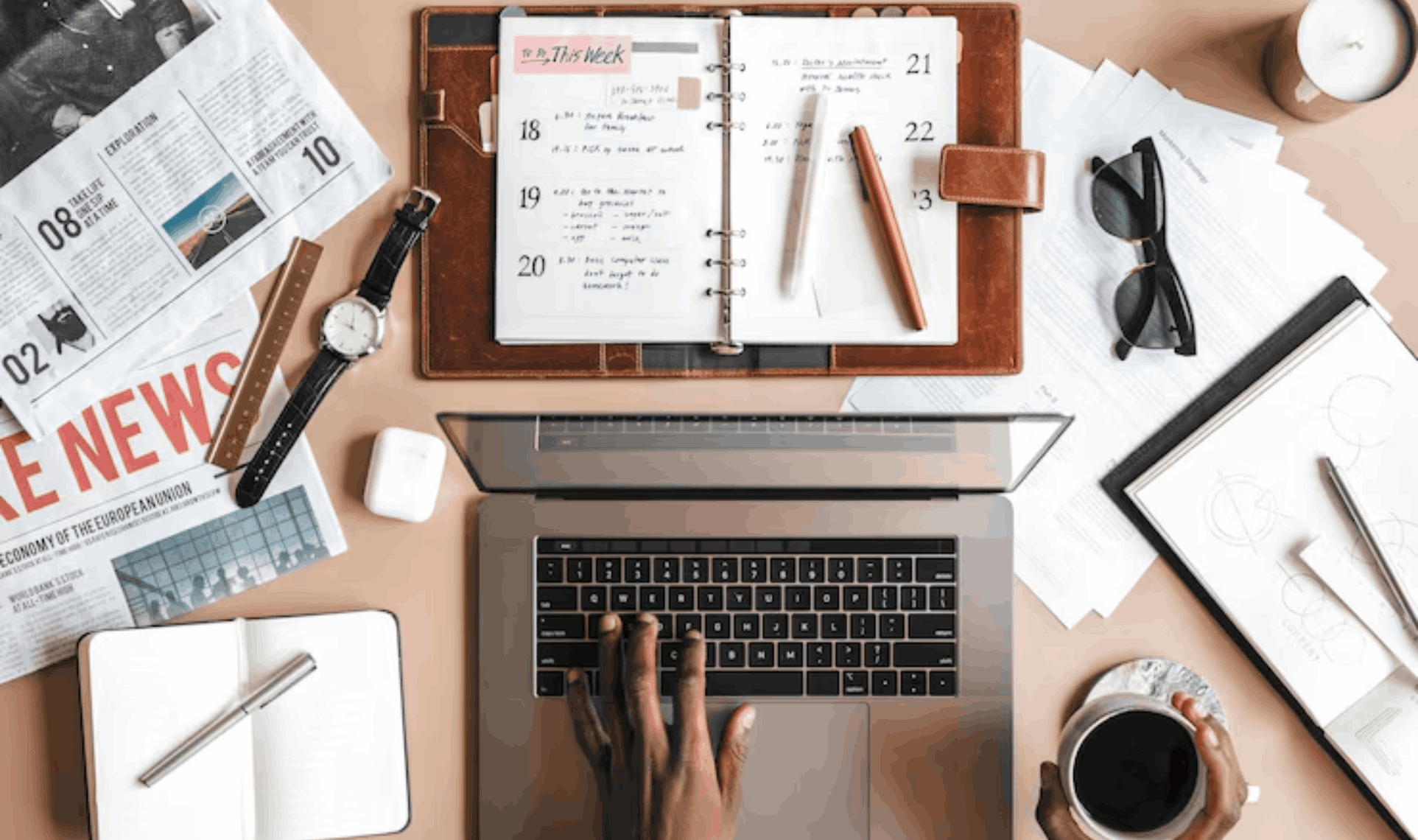 Workspace with laptop, planner, newspapers, and notebooks used for organizing Case Studies & Press Releases, with a person typing and holding a cup of coffee.