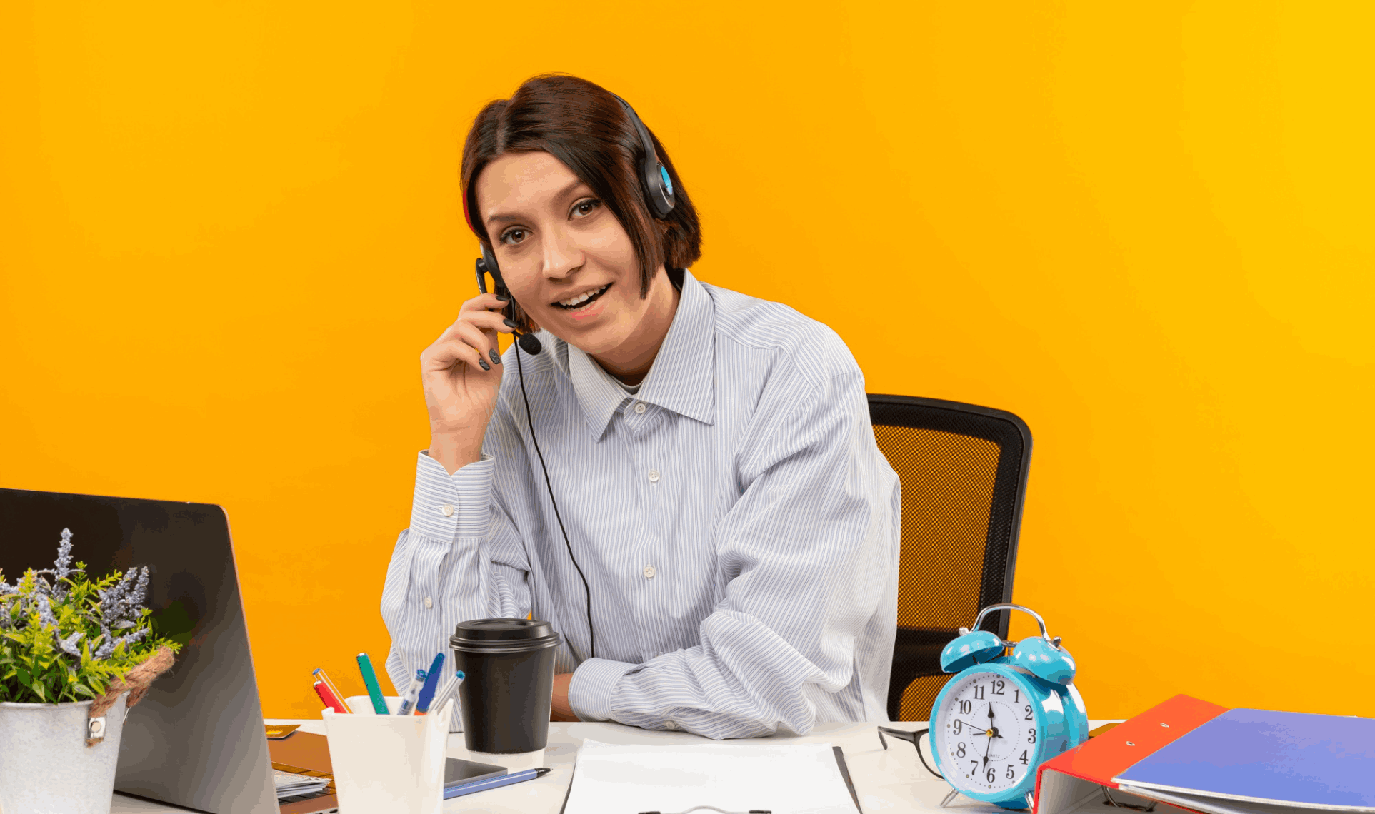 A smiling female customer service representative wearing a headset and holding a microphone, sitting at a desk with a laptop and coffee.