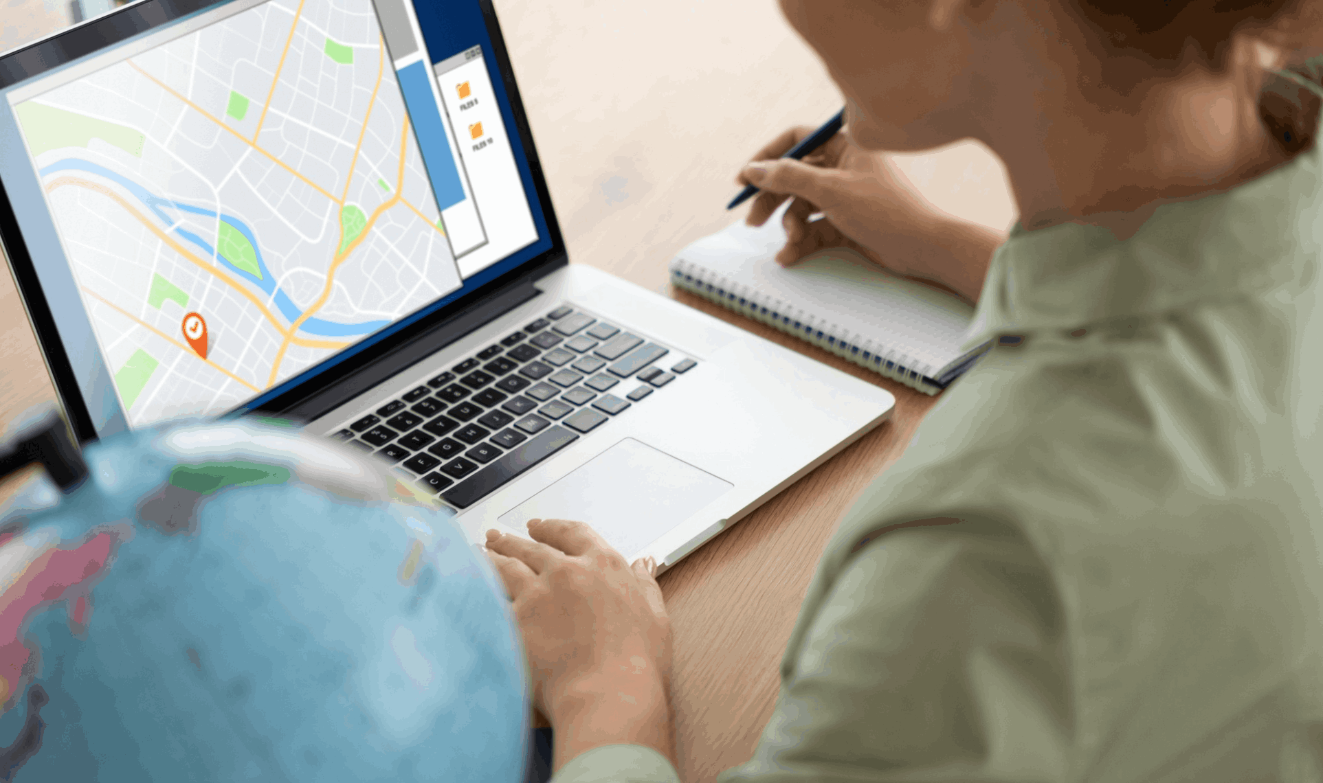 Person using laptop with online map open while taking notes, globe in foreground.