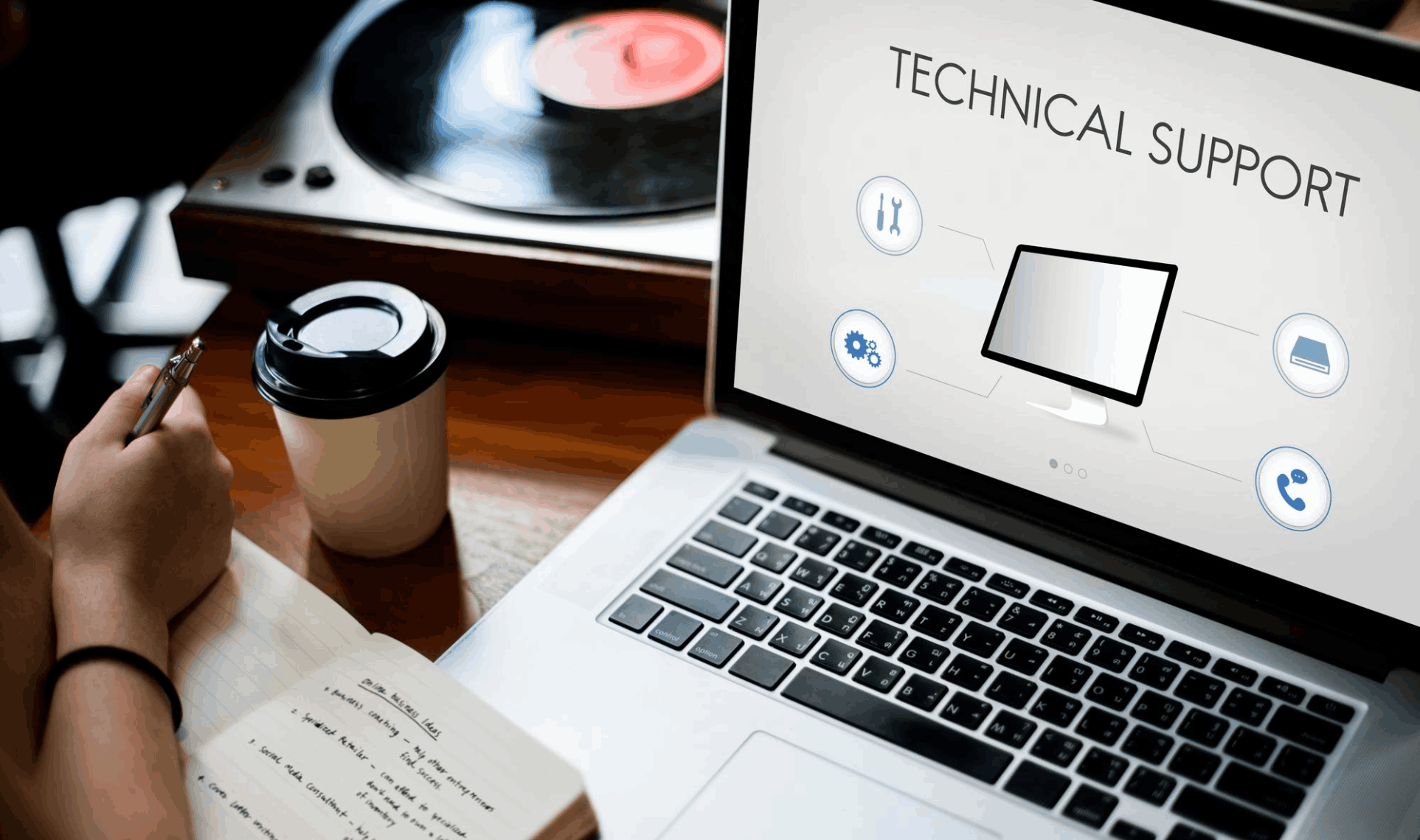 Person taking notes beside a laptop displaying a “Technical Support” screen, with a coffee cup and a vinyl record player in the background - Technical & Application Support.